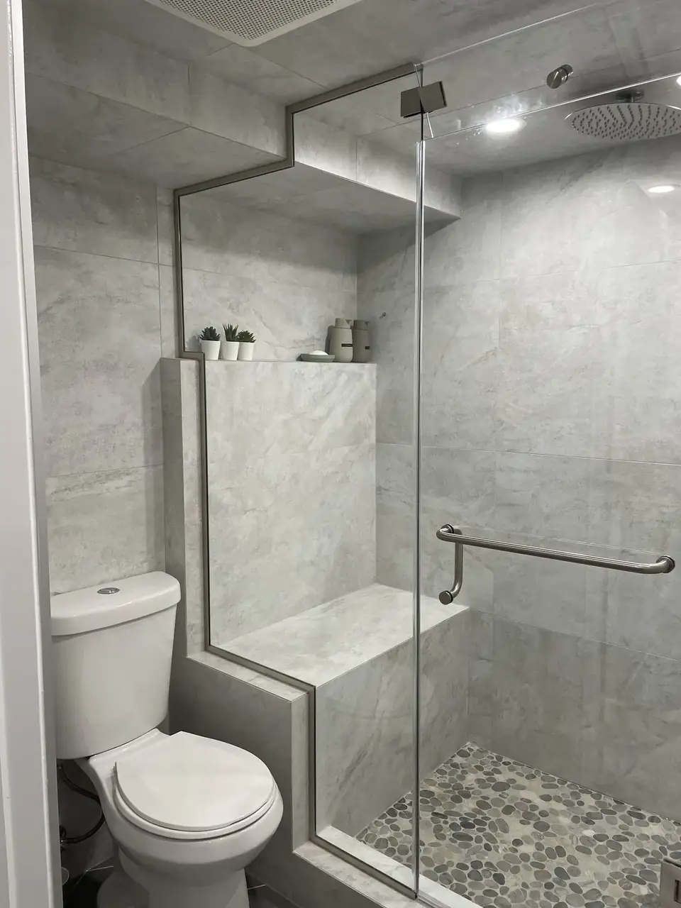 Walk-in shower with grey wall tile, a pebble floor, a glass panel and a grab bar