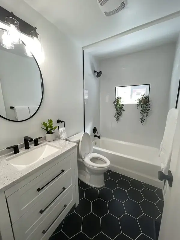 White vanity with a round mirror over a black hex tile floor, tub alongside